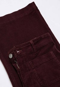 Pantalon en velours côtelé bordeaux foncé plié montrant les détails de la poche avant, du bouton et de la fermeture éclair sur fond blanc.
