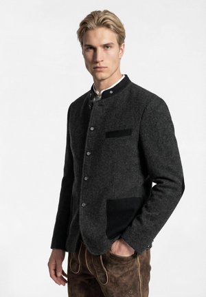 Junger Mann mit blondem Haar, der eine dunkelgraue Jacke mit Knöpfen und braune traditionelle Lederhosen trägt, mit einer Hand in der Tasche.