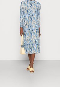 Blå blommönstrad klänning med långa ärmar, rund halsringning och midilängd. Matchas med gula sandaletter och en liten handväska.