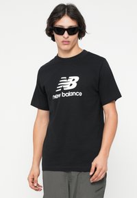 Juodas trumpas rankovėmis marškinėliai su apvaliu iškirpimu, kuriuose yra baltas "New Balance" logotipas. Audinys atrodo lygus, dizainas minimalistiškas.