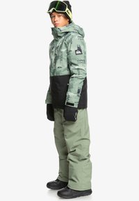 Zielona kurtka śnieżna z czarnymi akcentami, w geometryczne wzory, z przednim zamkiem, zestawiona z jasnobrązowymi spodniami śnieżnymi i czarnymi butami narciarskimi.