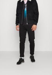 Chaqueta negra de pana con cuello de borrego blanco, combinada con una camiseta gráfica oscura y pantalones negros de corte slim. Zapatillas negras con calzado con estampado.