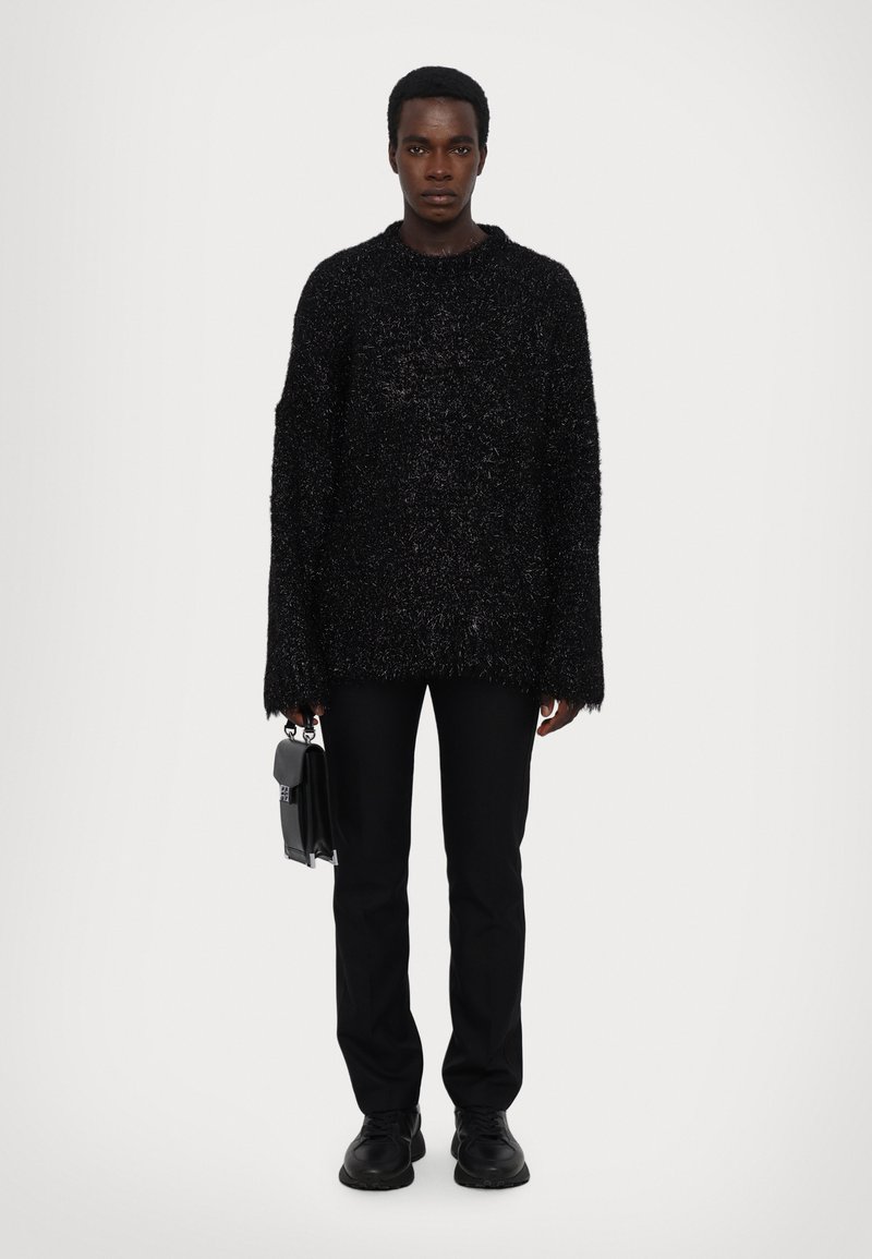 Pulover oversized, texturat, de culoare neagră, cu mâneci lungi, asortat cu pantaloni slim negri și pantofi negri robusti, ținând în mână o geantă mică neagră.
