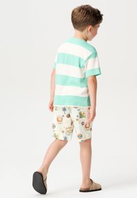 Gestreiftes Kurzarmhemd in Mintgrün und Weiß; gemusterte Shorts aus leichtem Stoff mit bunten Grafiken; beiger Slipper.