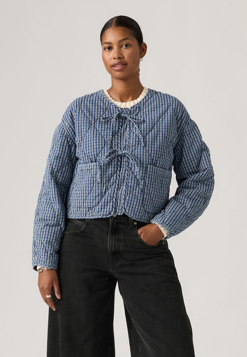 Γυναίκα που φοράει μπλε μπουφάν με επένδυση σε καρώ σχέδιο, με δέσιμο μπροστά, πάνω από εκρού τοπ, συνδυασμένο με ψηλόμεσα μαύρα τζιν παντελόνια με φαρδιά μπατζάκια.