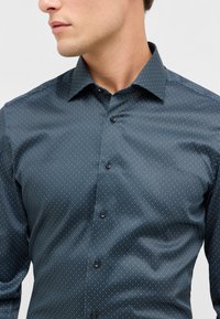 Donkerblauwe blouse met knopen, voorzien van een klein geometrisch patroon van witte en gele stippen, met een gestructureerde kraag en een gladde textuur.