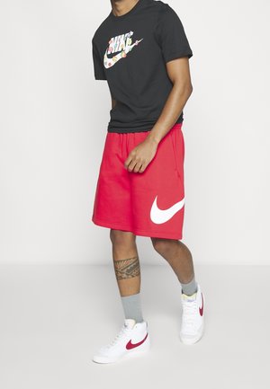 Mężczyzna noszący czarną koszulkę Nike z kolorowym logo, czerwone spodenki Nike z białym znakiem Swoosh, szare skarpetki i białe buty Nike z czerwonym znakiem Swoosh, stojący na białej podłodze.