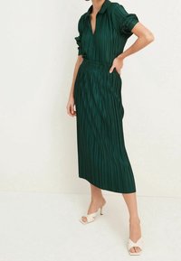 Robe midi plissée verte avec un col, une taille élastique et des manches à poignets ; associée à des sandales à talons à lanières de couleur claire.