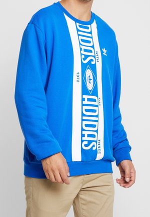 Mężczyzna noszący niebieską bluzę Adidas z białymi pionowymi paskami i napisem, w połączeniu z beżowymi spodniami.
