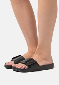 Schwarze Rutsch-Sandalen verfügen über einen breiten Riemen mit einer großen Schnalle, eine flache Sohle und ein offenes Zehendesign, das eine glatte Textur und einen minimalistischen Stil zur Geltung bringt.