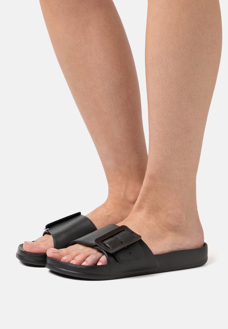Schwarze Rutsch-Sandalen verfügen über einen breiten Riemen mit einer großen Schnalle, eine flache Sohle und ein offenes Zehendesign, das eine glatte Textur und einen minimalistischen Stil zur Geltung bringt.