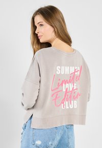 Grå kort sweatshirt laget av mykt stoff med hvit og rosa tekst på baksiden som sier "SUMMER Limited Edition CLUB." Slitte blå jeans er synlige.