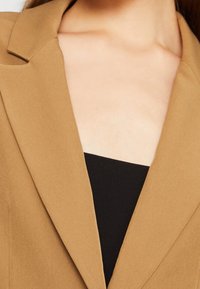 Hnědý blejzr s klopovým límcem, hladkým materiálem a střihaným střihem, nošený přes černý top, zdůrazňující límec a výstřih.