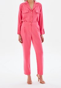 Ensemble deux pièces rose en satin comprenant une chemise boutonnée avec deux poches poitrine et un pantalon à jambes droites. Texture lisse et coupe ajustée.