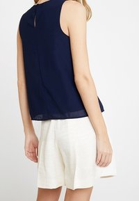 Haut sans manches bleu marine avec une ouverture en forme de goutte dans le dos, associé à un short crème texturé. Le haut présente une finition lisse et une coupe décontractée.