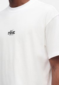 Λευκό κοντομάνικο T-shirt με μικρό μαύρο λογότυπο Nike και κείμενο "sportswear" στο στήθος, φορεμένο από άτομο με σκουρόχρωμο δέρμα.
