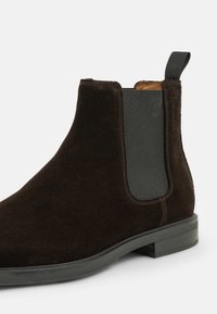 Bottines Chelsea en suede marron avec des panneaux latéraux élastiques, une languette et une semelle en caoutchouc. Texture lisse avec des détails de couture.