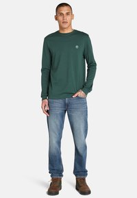 Langärmliges grünes Hemd mit Rundhalsausschnitt und Logo auf der Brust, kombiniert mit blauen Jeans und braunen Schnürstiefeln.