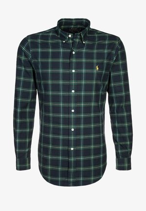 Uomo con camicia a quadri verde scuro e jeans neri che tiene in una mano una felpa grigia mentre cammina.