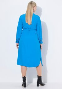 Sininen pitkähihaisen mekko, jossa on vyö, sivurei'ät ja nappisuljinteet. Liukas pinta ja virtaava muotoilu, yhdistettynä kiiltäviin mustiin nilkkasaappaisiin.