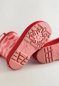 Rote Gummistiefel mit Herzenmuster und strukturierten Sohlen mit verspieltem Spritzdesign. Weiches rosa Innenfutter und Akzente.