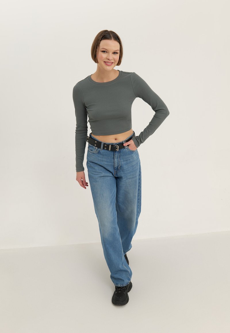 Νεαρή γυναίκα που χαμογελά, φοράει γκρι crop top με μακριά μανίκια, άνετο μπλε τζιν με μαύρη ζώνη και μαύρα αθλητικά παπούτσια, στέκεται μπροστά σε λευκό φόντο.