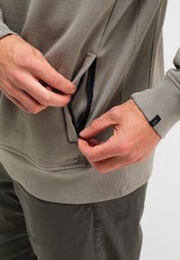 Heller grauer Sweatshirt mit einem Reißverschlussfach, gerippten Bündchen und einer weichen Textur. Hände zeigen die Funktionalität der Tasche.