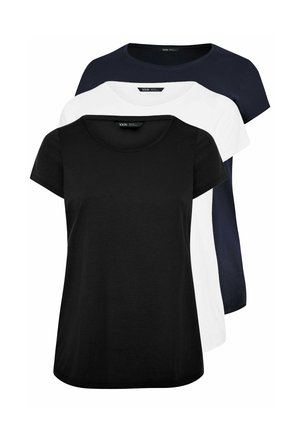 Blondynka stoi na tle gładkiej ściany, ubrana w czarną bluzkę z krótkim rękawem, niebieskie dopasowane jeansy i białe sneakersy.