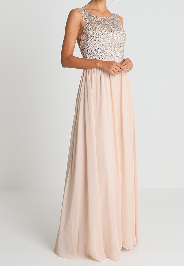 Femme portant une robe longue sans manches avec un corsage à sequins et une jupe fluide rose clair, debout, les mains jointes devant.