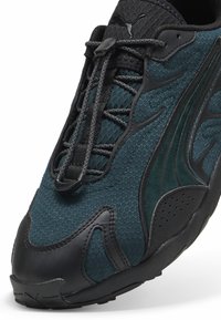Sneakers Puma z ciemnozielonym teksturowanym siateczkowym wierzchem, czarnymi akcentami, heksagonalnym wzorem i czarną gumową podeszwą. Wyposażone w bezpieczny system sznurowania.