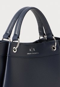 Borsa in sintetico testurizzato blu navy con accessori metallici rotondi, doppio manico superiore e logo impresso 'A|X ARMANI EXCHANGE' sul lato.