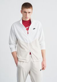 Chaqueta con cremallera de Nike en tela blanca y crema, con un cuello alto y un logo negro en el pecho. Combinada con pantalones jogger crema.