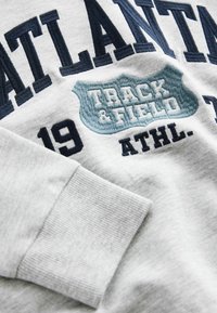 Szary sweter z niebieskim haftowanym napisem "ATLANTA", "TRACK & FIELD" oraz numerem "19". Posiada ściśnięte mankiety i miękką teksturę.