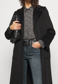 Abrigo largo negro sobre una camisa gris con estampado, combinado con jeans oscuros. Sostiene un clutch negro texturizado. Presenta accesorios de hardware plateado en el cinturón.