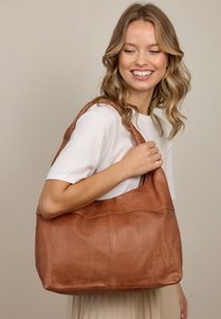 Brun læder tote bag med en blød, struktureret overflade, der har en bred åbning og et vævet skulderrem. Glatte sider og minimalistisk design.