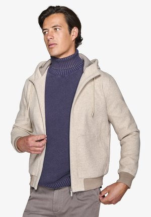 Helle beigefarbene Kapuzenjacke mit weicher Textur, mit einem Reißverschluss vorne, Kordelzug und zwei Fronttaschen. Getragen über einem dunkelblauen, gerippten Rollkragenpullover.