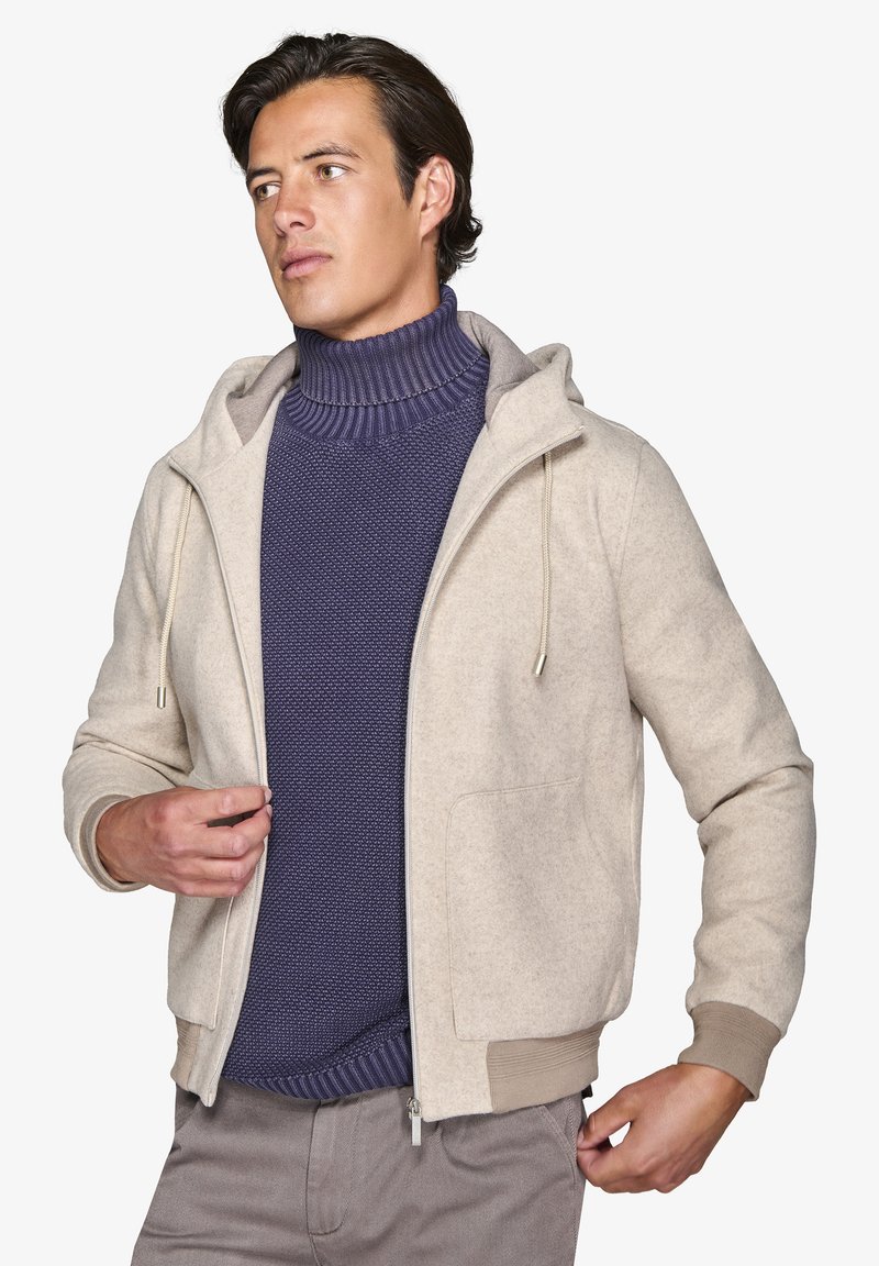 Lys beige hættetrøjejakke med en blød tekstur, der har lynlås foran, snor og to forlommer. Båret over en mørkeblå, ribbet turtleneck.