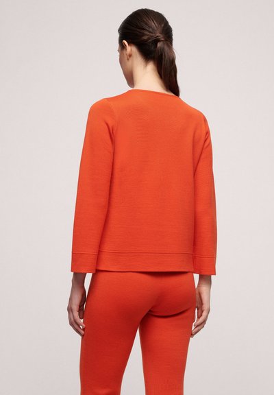 Oranges Stricktop mit Dreiviertelärmeln und gerader Saum, kombiniert mit orangen, figurbetonten Hosen. Glatte Textur, dezente Musterung.