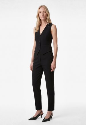 Blonde Frau trägt eine taillierte schwarze ärmellose Weste mit Knöpfen, passende schlanke schwarze Hosen und schwarze, niedrig-absätzige spitze Schuhe.