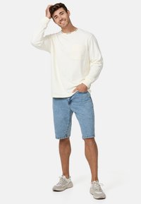 T-shirt à manches longues crème clair avec une poche sur la poitrine, associé à un short en denim bleu clair. Le mannequin porte des baskets grises.