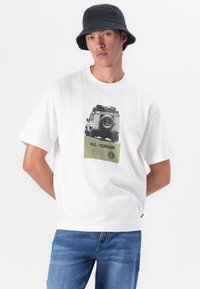 Camiseta blanca de algodón con un gráfico de un vehículo todoterreno y el texto "TODO - TERRENO" con un acento verde en la parte inferior. Combinada con jeans azules.