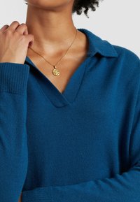 Blauwe gebreide trui met een V-hals en kraag, met ribbelmanchetten. Een gouden hangersnoer met een ronde charm hangt op de halslijn.