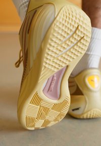 Sneaker giallo chiaro con suola testurizzata, accenti rosa e motivi sottili. Presenta un tallone imbottito e un design a strati per un maggiore supporto.