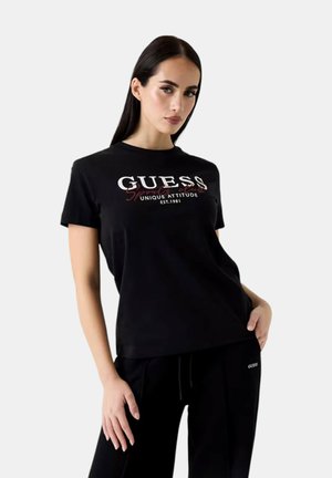 Donna con lunghi capelli scuri che indossa una T-shirt Guess nera con scritte bianche e rosse, abbinata a pantaloni Guess neri, posando con una mano in tasca.