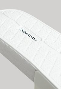 Suela de sandalia de goma blanca con un patrón texturizado de formas rectangulares. La marca "SUPERDRY" está grabada en el centro.