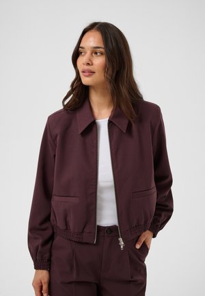 Femme aux cheveux bruns mi-longs, portant une veste zippée violet foncé et un pantalon assorti par-dessus un haut blanc, regardant légèrement sur le côté.