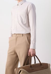 Raidallinen beige nappipaita, jossa on rintatasku, yhdistetty vaaleanruskeisiin räätälöityihin housuihin. Kädessä ruskea ja beige nahkalaukku.