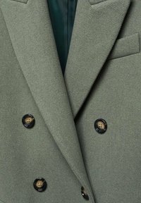 Manteau en laine vert olive à double boutonnage avec revers à pointe, quatre boutons noirs, et une doublure intérieure vert foncé visible au col.