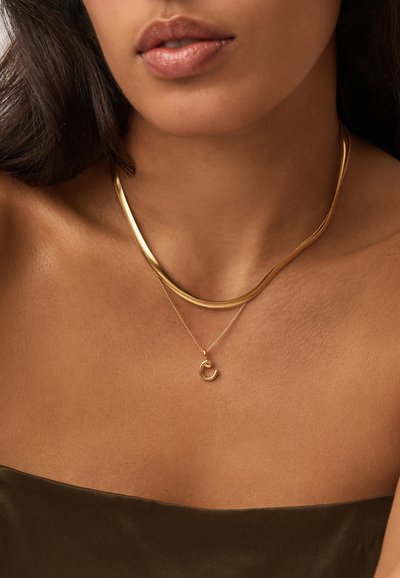 Γυναίκα με μέτριο τόνο δέρματος φοράει δύο χρυσά κολιέ, ένα επίπεδη αλυσίδα και ένα λεπτό αλυσίδα με ένα μικρό μενταγιόν σε σχήμα ημισελήνου, ντυμένη με σκούρο καφέ τοπ.