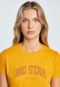 Geltona medvilninė marškinėliai su trumpomis rankovėmis, su dideliu nusidėvėjusiu spaudiniu, ant kurio parašyta "BIG STAR JEANS" tamsesne spalva.